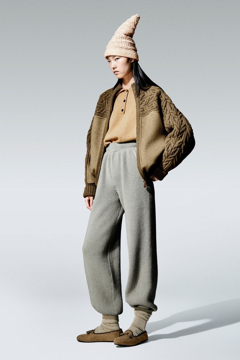 Asian woman poses in a knit hat, brown cable-knit jacket, tan shirt, and gray knit pants against a neutral gray background.
