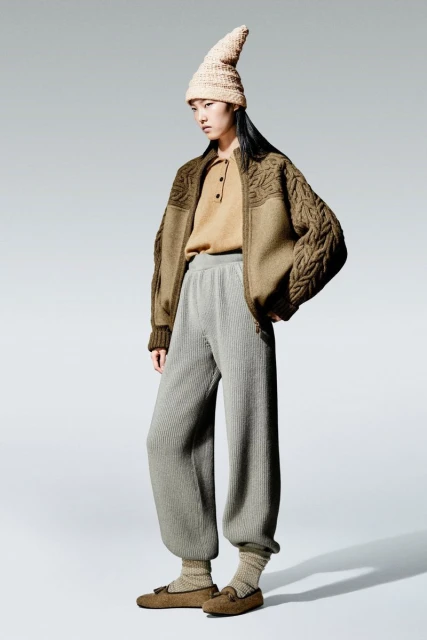 Asian woman poses in a knit hat, brown cable-knit jacket, tan shirt, and gray knit pants against a neutral gray background.