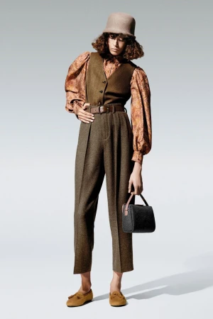 A fashion model stands against a plain grey background wearing a felt bucket hat, paisley shirt, olive wool vest, trousers, and loafers.