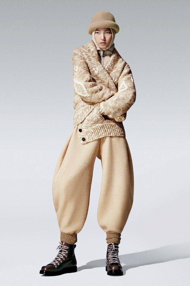 Person posing in beige sweater, oversized pants, boots, and beanie in a full shot against a plain background in a studio setting.