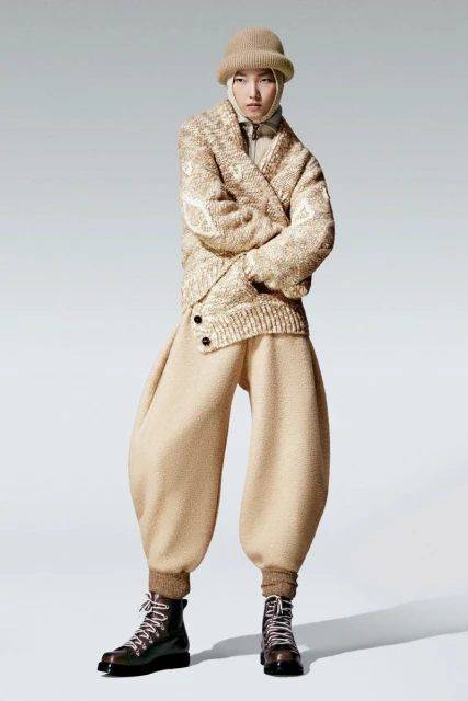 Person posing in beige sweater, oversized pants, boots, and beanie in a full shot against a plain background in a studio setting.