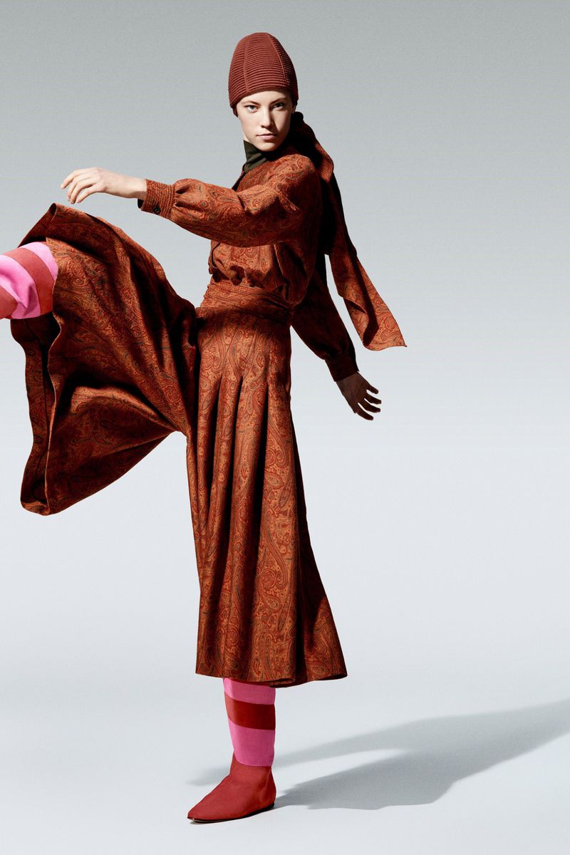 A woman in a patterned brown outfit with a matching hat poses dynamically against a light grey background with pink-striped socks.