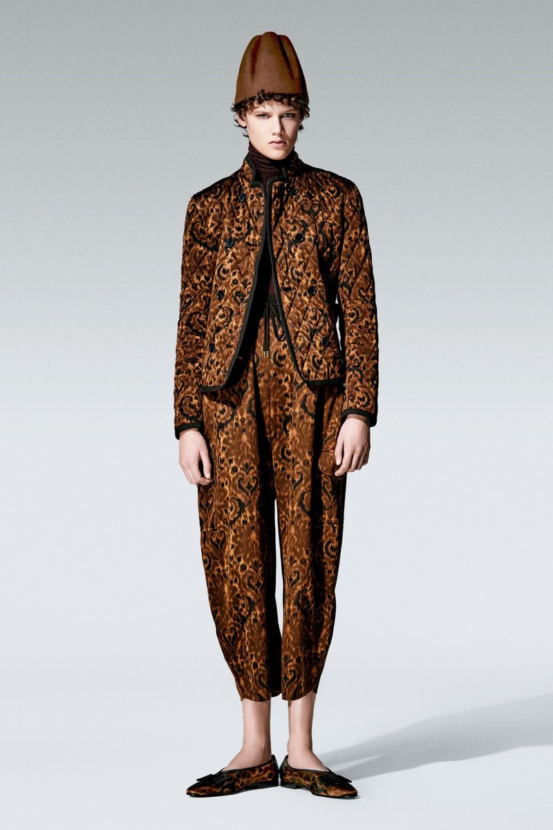 Full shot of a person with curly hair wearing a brown patterned jacket, pants, shoes, and a hat in a studio against a grey background.
