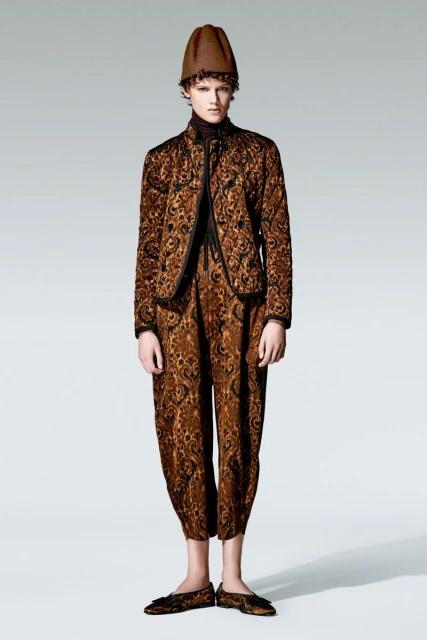 Full shot of a person with curly hair wearing a brown patterned jacket, pants, shoes, and a hat in a studio against a grey background.