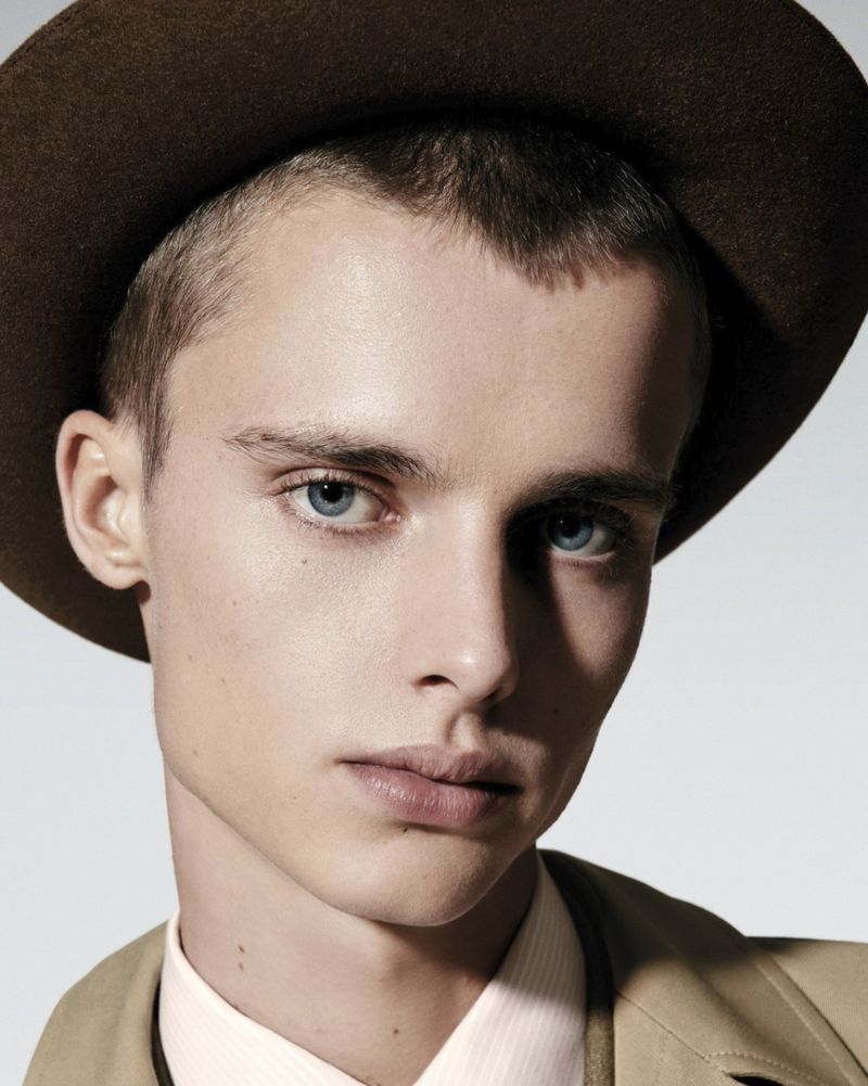 Close-up portrait of an adult male with light skin, blue eyes, and short hair, wearing a brown hat and trench coat against a light background.