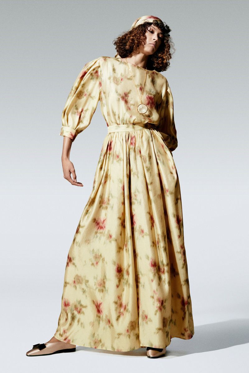 Full-length portrait of a female fashion model wearing a long, floral-print dress with matching headband in a light studio.