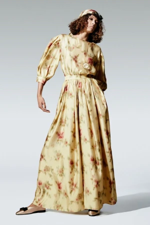Full-length portrait of a female fashion model wearing a long, floral-print dress with matching headband in a light studio.
