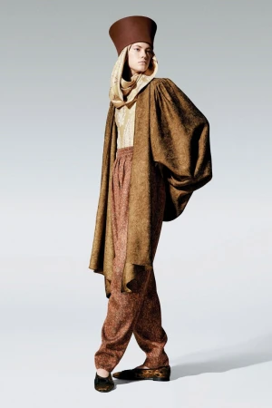 Model wearing a tall brown cylindrical hat, patterned scarf, and oversized coat against a plain white background