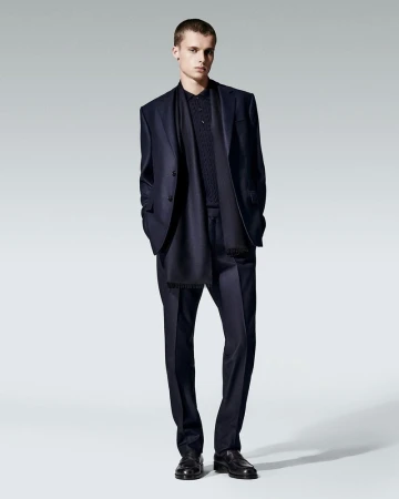 Man wearing a dark navy suit jacket, matching trousers, polo shirt, and a scarf against a neutral grey studio background.