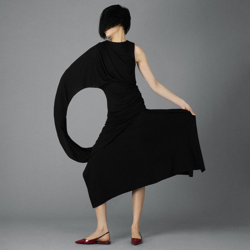 A woman in a black dress with a large circular cutout stands against a grey backdrop, presenting a modern and stylish fashion statement.
