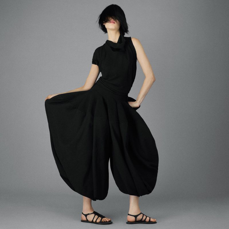 Adult female poses in wide-leg black pants and a high-necked sleeveless top against a neutral gray background, showcasing fashion style.