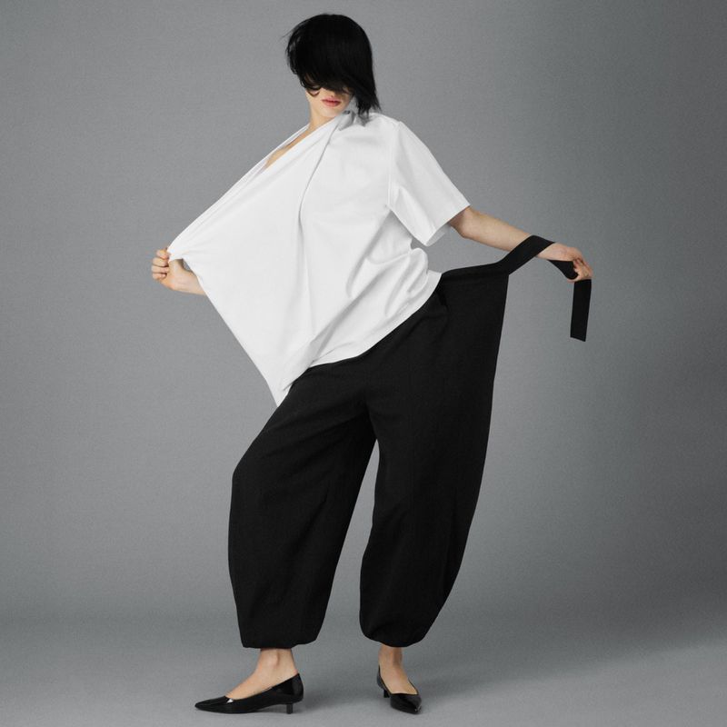 A woman in a white kimono-style top and black balloon pants poses against a neutral background, showcasing a modern, chic fashion look.