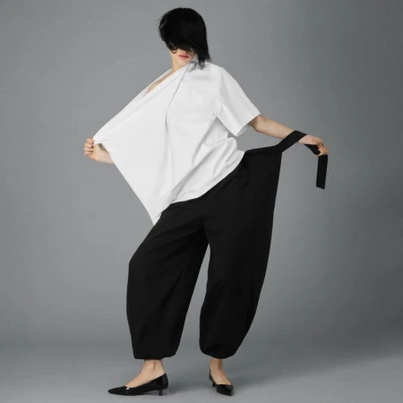 A woman in a white kimono-style top and black balloon pants poses against a neutral background, showcasing a modern, chic fashion look.