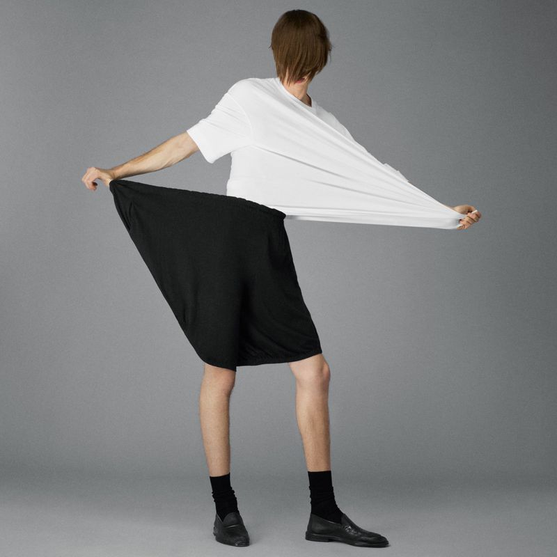 Person stretching an oversized white T-shirt and black fabric, standing in black socks and dress shoes on a gray backdrop in a studio.