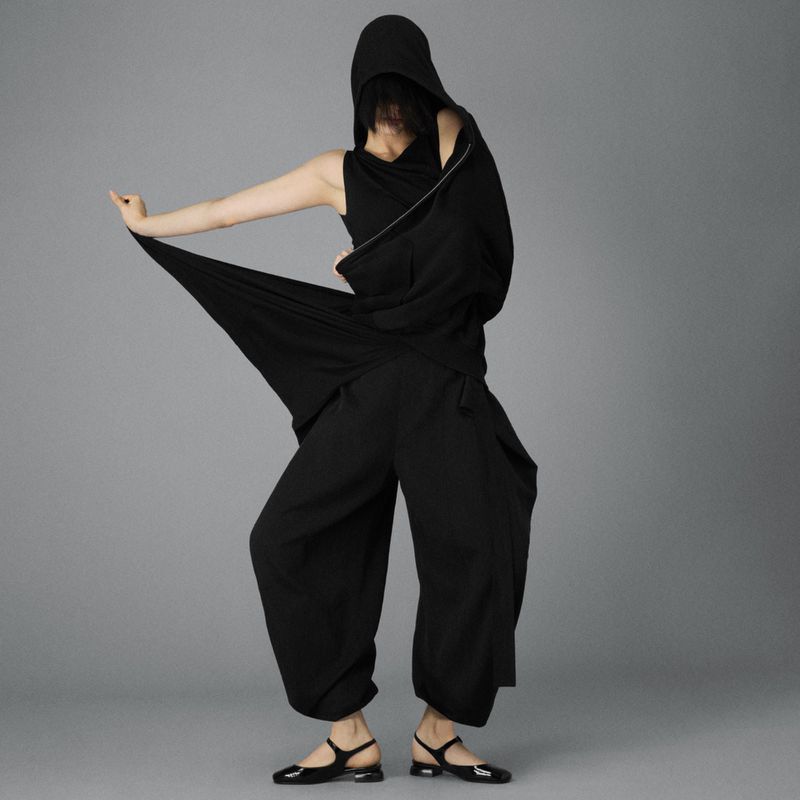 Woman wearing an avant-garde black hooded outfit strikes a dramatic pose in a grey studio, showcasing modern fashion design and style.