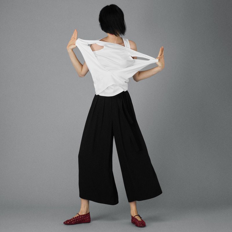 Back view of a person wearing a white tank top and black wide-leg pants, posing against a gray background. Fashion shot.