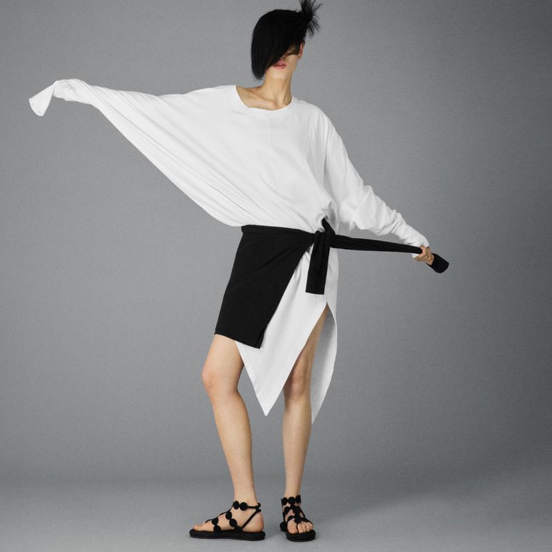 A stylish person in a white long-sleeve top and black wrap skirt poses in a studio with hands outstretched, wearing a black hat and sandals.