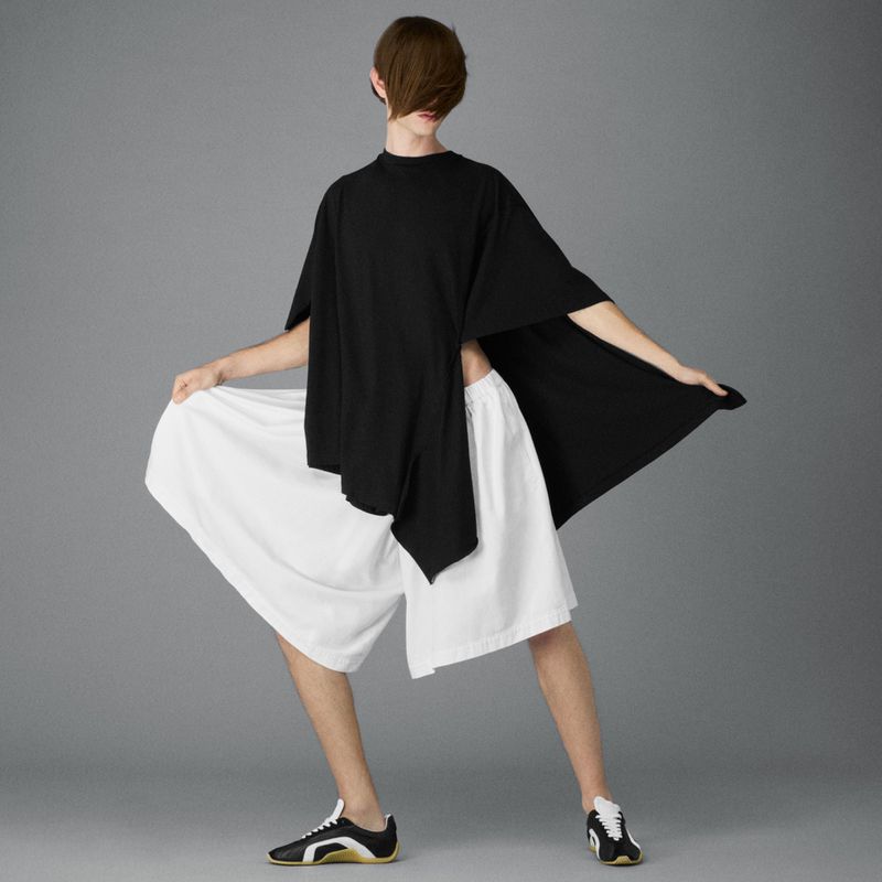 Person posing in a black top and white shorts against a grey background, showcasing a relaxed fashion style with visible tennis shoes.