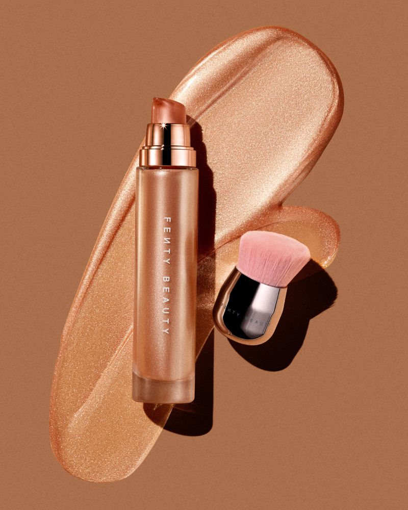 Fenty Beauty face and body luminizer next to face brush against tan backdrop, lit with a strong key light for high shine.