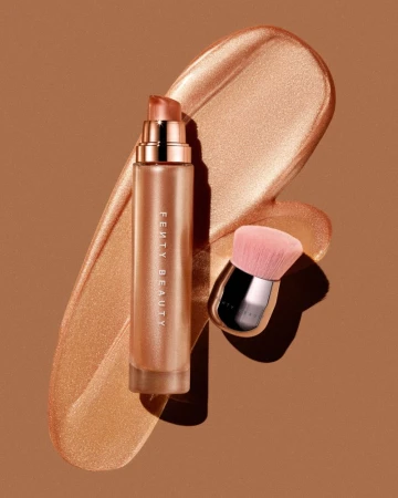 Fenty Beauty face and body luminizer next to face brush against tan backdrop, lit with a strong key light for high shine.