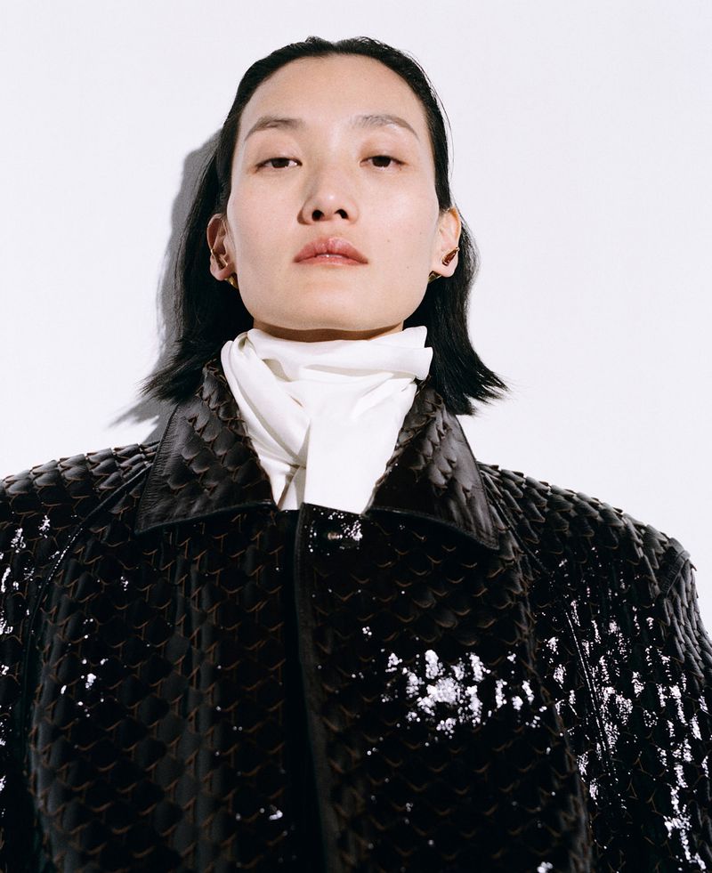 Close-up portrait of an adult woman with black hair in a shiny, textured black jacket with white turtleneck against a white backdrop.