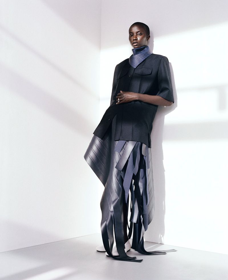 Fashionable adult female model poses in a black, short-sleeved jacket and avant-garde skirt with rubber elements in a photo studio.