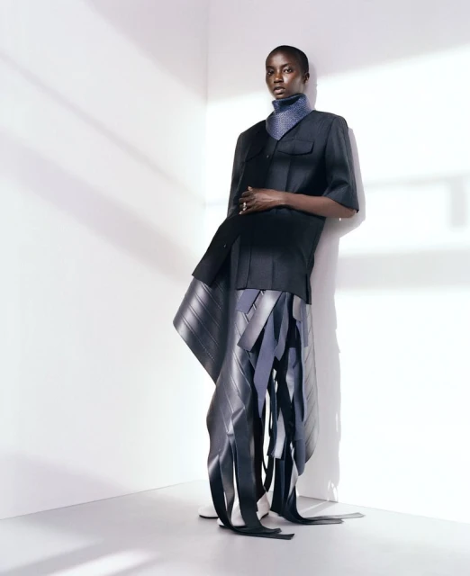 Fashionable adult female model poses in a black, short-sleeved jacket and avant-garde skirt with rubber elements in a photo studio.