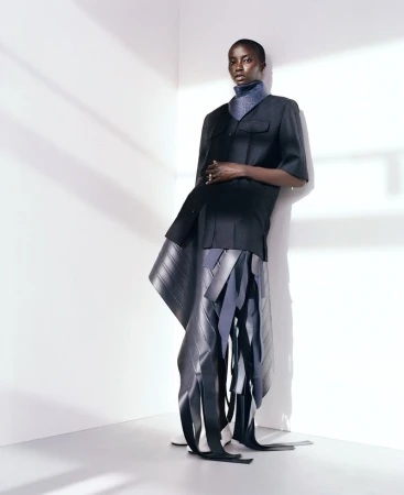 Fashionable adult female model poses in a black, short-sleeved jacket and avant-garde skirt with rubber elements in a photo studio.