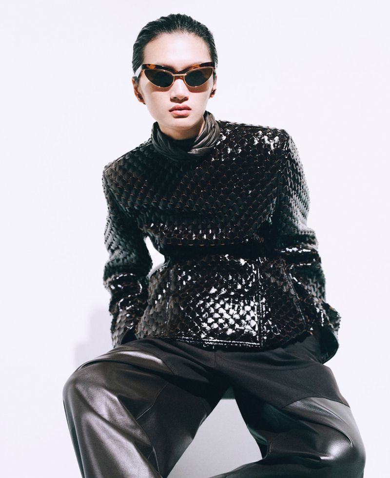 A person with slicked-back hair wears cat-eye sunglasses and a dark textured woven jacket against a stark white studio background.