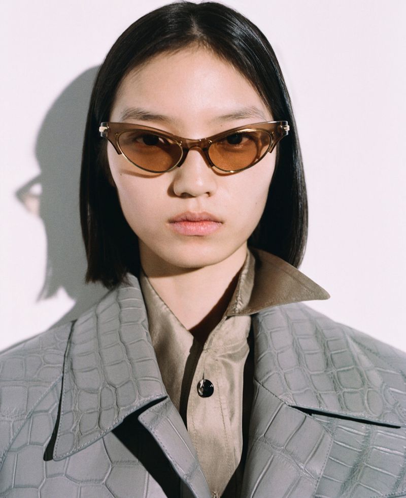 Portrait of a young Asian woman with dark hair and sunglasses, wearing a collared shirt and reptile-texture jacket against a white backdrop.