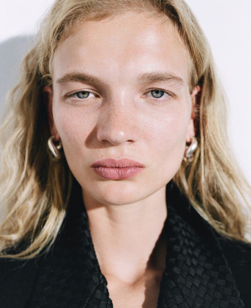 Close-up portrait of a fair-skinned woman with blonde wavy hair, light blue eyes, and silver hoop earrings, wearing a black jacket.