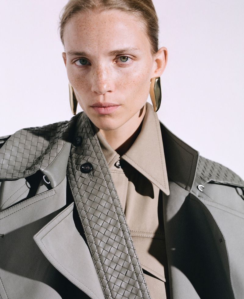 Portrait of a fair-skinned young woman with freckles, wearing a woven trench coat and large earrings, looking directly at the camera in a studio setting.