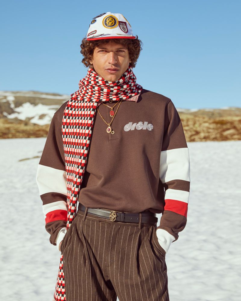 Model wearing a brown polo sweatshirt, patterned scarf, and cap against a snowy mountain landscape for Drole De Monsieur