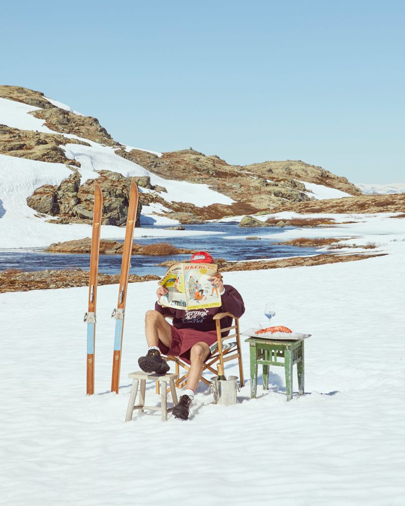 Man reads magazine in snowy mountain landscape, seated in director's chair next to skis. Clear blue sky, bright outdoor light.