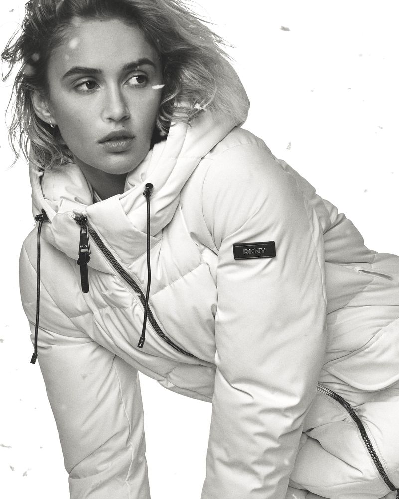 A light-skinned female model poses in a three-quarter shot, wearing a winter puffer coat, indoors against a white background with snow.