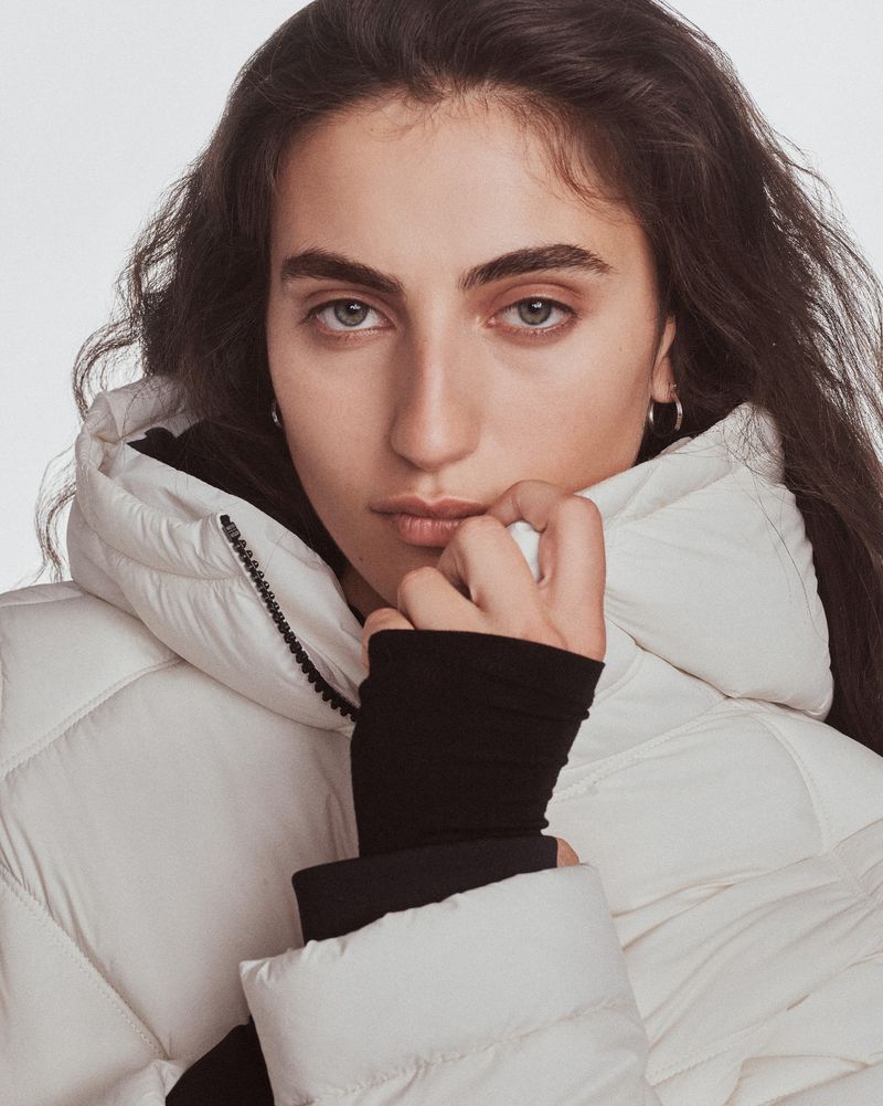 Close-up portrait of a brunette woman wearing a puffy white jacket with a black top underneath, posing in front of a white backdrop.