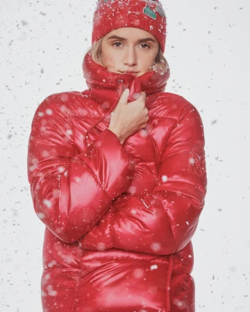 A blond woman wearing a red puffer coat and a matching red beanie in a studio setting with falling snow effect. Winter fashion.
