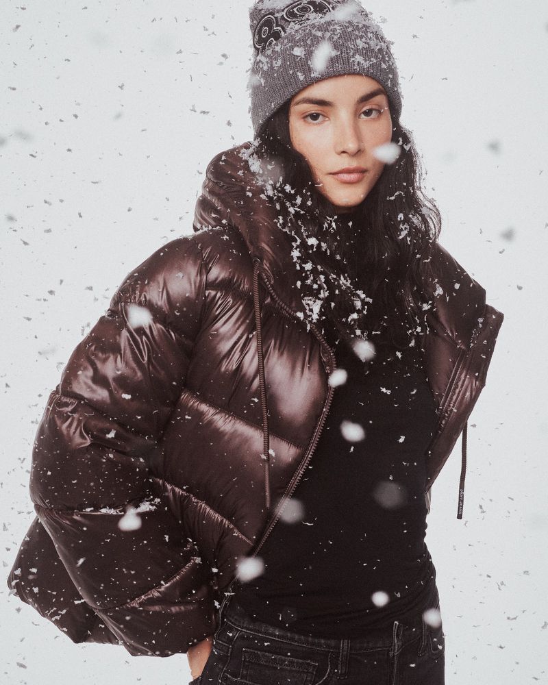 A woman in a grey knit hat and puffy brown jacket stands against a white backdrop with snow falling in the foreground.