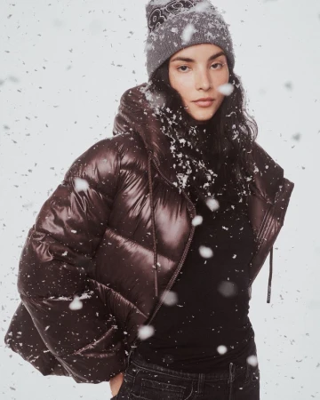 A woman in a grey knit hat and puffy brown jacket stands against a white backdrop with snow falling in the foreground.
