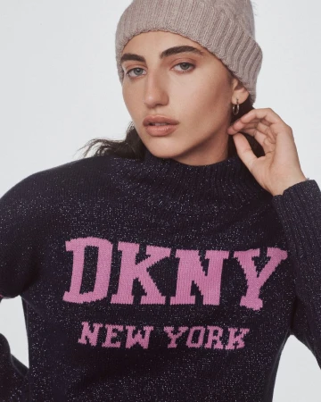 A young adult woman with dark hair and a beige beanie poses indoors wearing a blue DKNY sweater with pink text, medium shot.