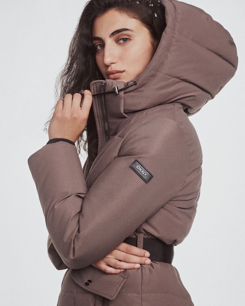 An adult woman in a hooded, belted, taupe puffer jacket looks slightly away from the camera in a brightly lit studio shot.