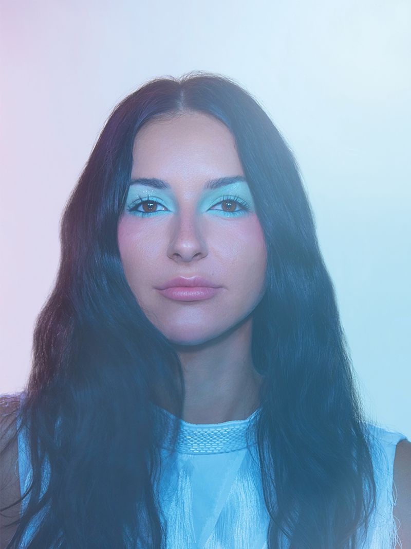 Portrait of a young woman with long dark hair and blue eye makeup, posing against a light blue and pink gradient background.