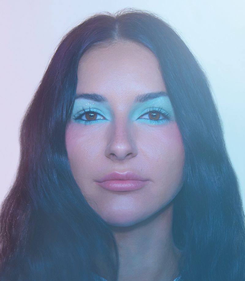Close-up portrait of a woman with long dark hair and fair skin wearing turquoise eye makeup and pink blush in a light-filled indoor space.