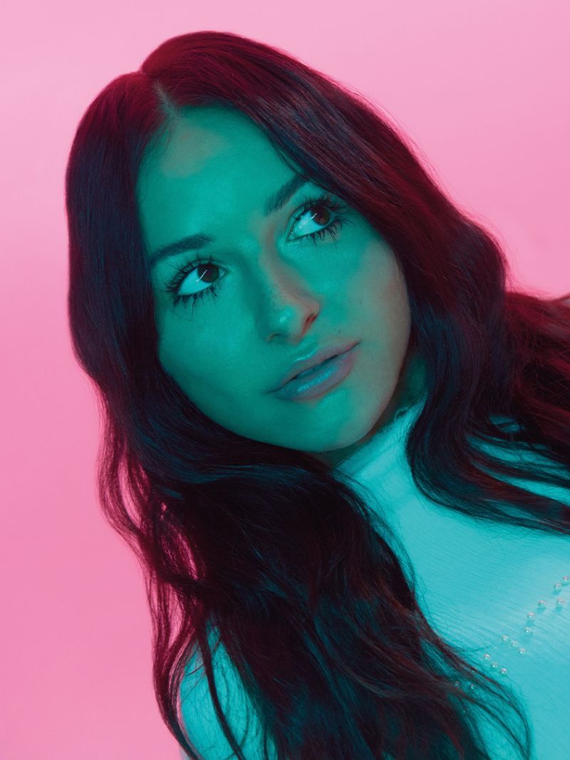 Portrait of a young adult female with dark wavy hair looking up, lit by neon teal and pink lights in a photo studio setting.