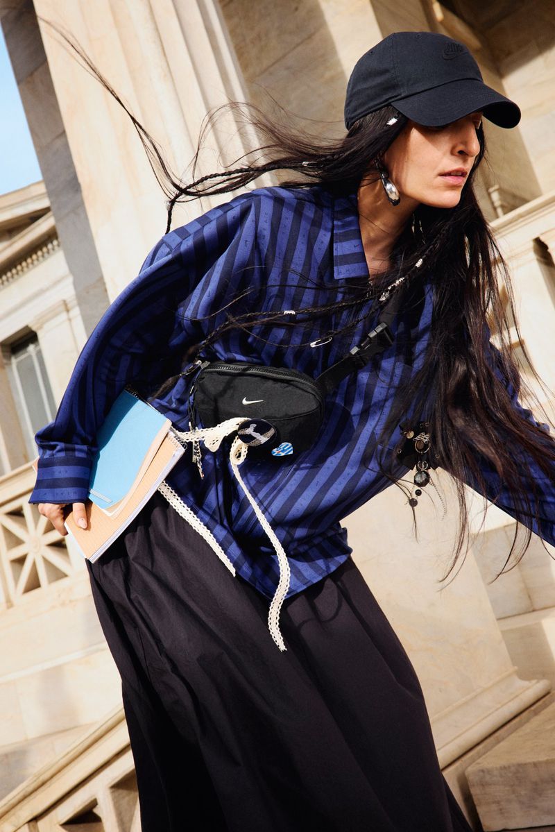 Fashion editorial photo of a woman in a blue striped shirt, black skirt and cap, walking on stairs with a small Nike bag and blue notebooks.