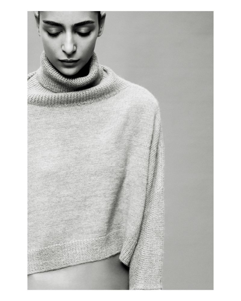 Monochrome portrait of a woman with short dark hair looking down, wearing a light grey wool turtleneck sweater on a grey background.