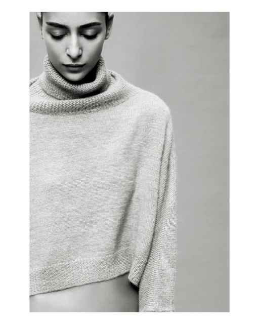 Monochrome portrait of a woman with short dark hair looking down, wearing a light grey wool turtleneck sweater on a grey background.