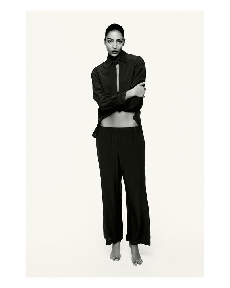 Portrait of a slender adult female wearing a black button-down shirt and loose pants against a plain white backdrop in a studio.