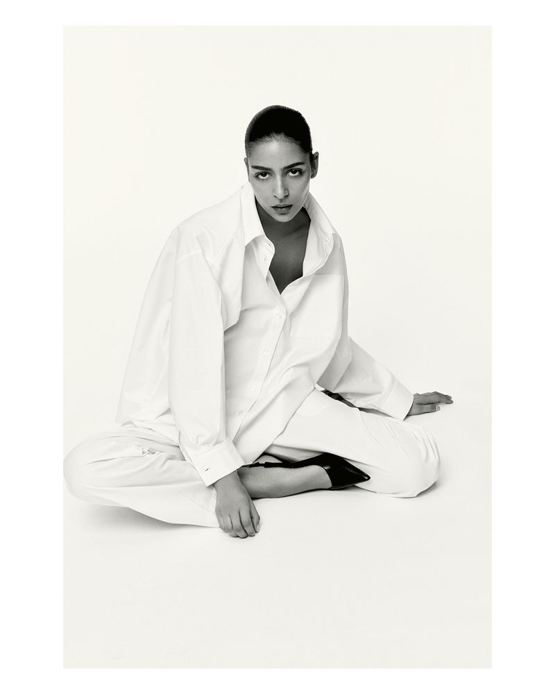 Black and white portrait of a young adult female model sitting on the floor wearing an oversized white shirt and black high heels.