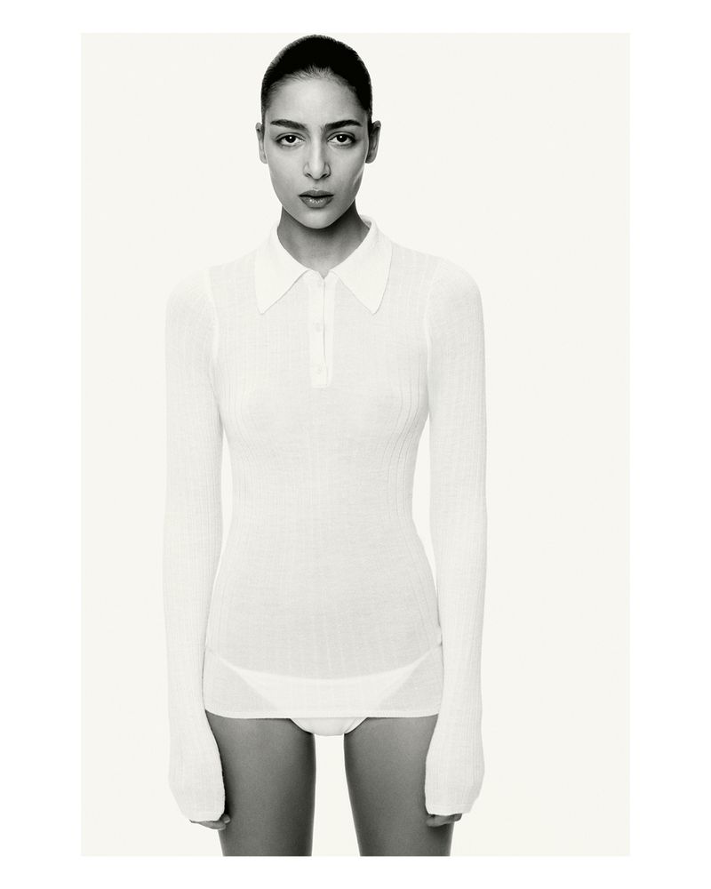 Studio portrait of an adult female fashion model wearing a white long-sleeved polo shirt and underwear against a bright neutral backdrop.